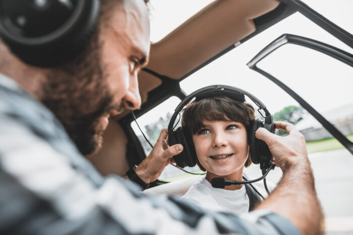 Optimistic child talking with glad pilot indoor Satisfied bearded man putting modern headsets on cheerful boy while communicating with him. They sitting in modern helicopter inside